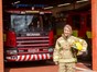 Female firefighter stood holding her helmet in front of an appliance bay with a fire engine parked inside